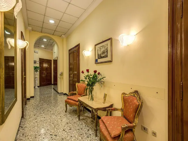 A hallway with a terrazzo floor, two gilded armchairs with red upholstery, a marble-topped side table holding a vase of red roses, and a framed print on the wall.