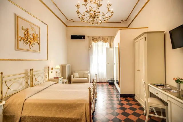 A bedroom with a brass-framed double bed, a gold relief panel on the wall, a chandelier, a checkered tile floor, an armchair by the window, a desk and wardrobe, and a wall-mounted television.