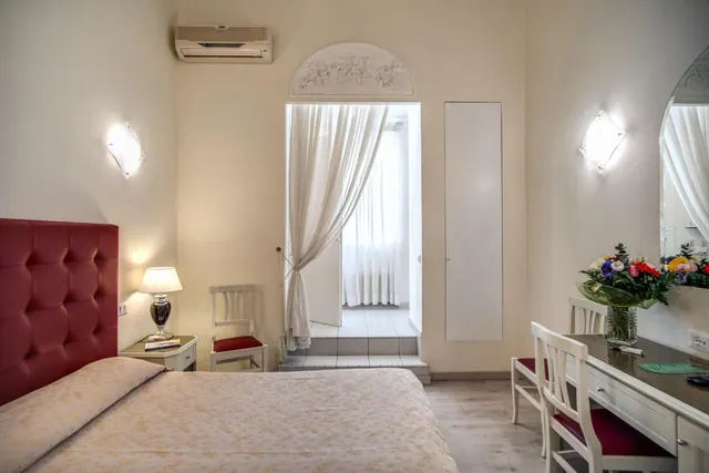 A bedroom with a red tufted headboard, a double bed, a bedside lamp, a white desk with a flower arrangement, and a doorway opening onto a tiled terrace.