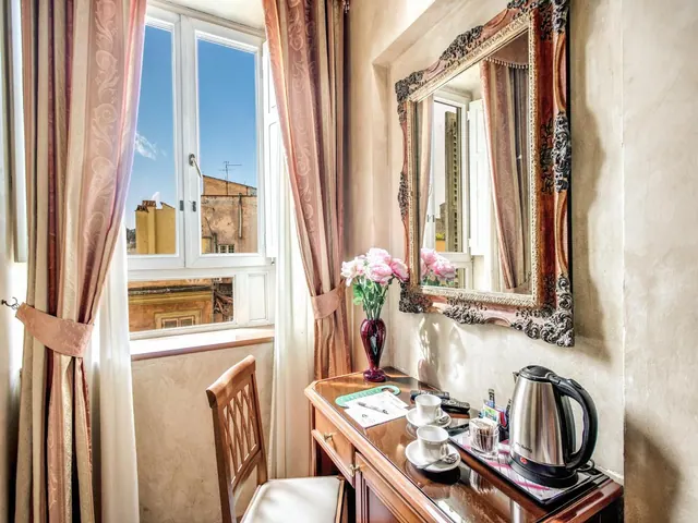 A wooden desk with a framed mirror, a vase of pink flowers, cups on saucers and an electric kettle stands beside a window with pink curtains that looks out onto neighbouring buildings and blue sky.