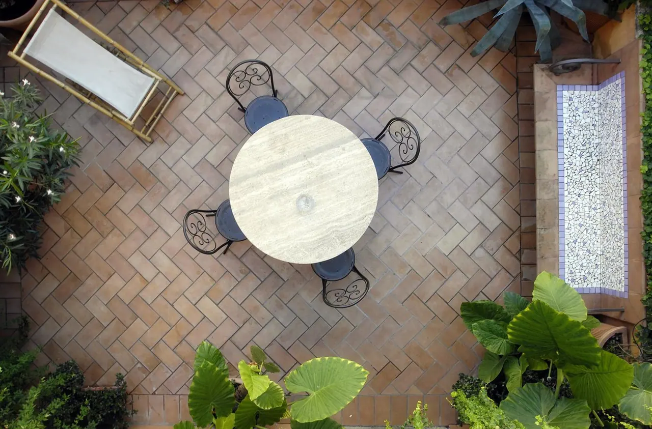 Overhead view of a brick-paved courtyard with a round stone table, four wrought iron chairs, a bamboo lounger, a small mosaic pool and surrounding plants.