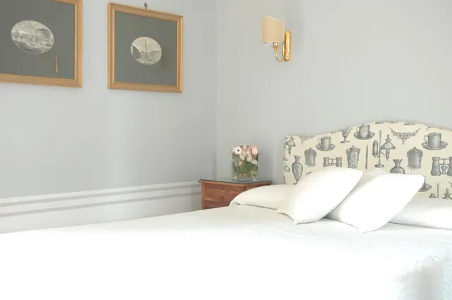 A bedroom with a white bed, a patterned upholstered headboard, two framed prints, a wall sconce and a wooden nightstand holding a small vase of flowers.