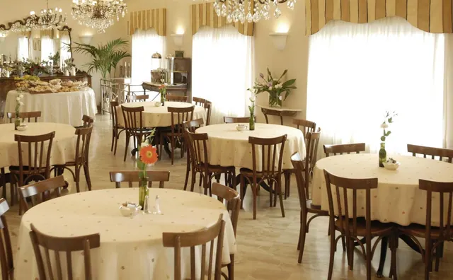 A dining room with round tables set with cream tablecloths, wooden bentwood chairs, chandeliers, and tall windows with white curtains.