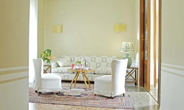 A living area with a patterned sofa, two white armchairs, a round wooden coffee table holding a tea service, and a patterned rug on a terrazzo floor.
