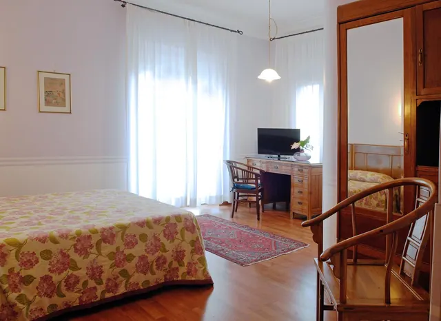 A bedroom with a double bed under a floral cover, a wooden desk with a television and chair, a wooden wardrobe with a mirror door, and white curtains over a window.