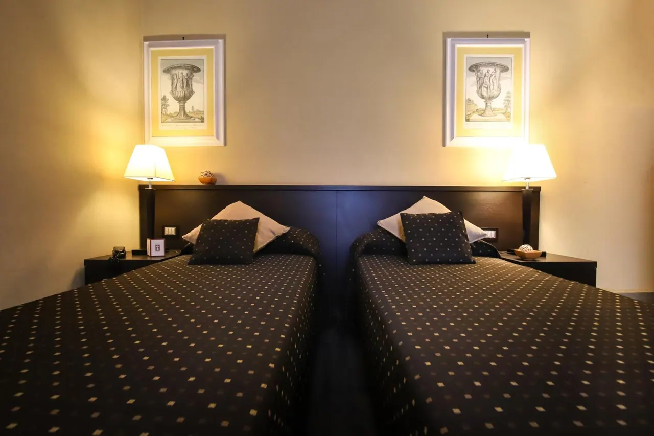 Two single beds with dark patterned bedspreads sit side by side under a dark headboard, with bedside lamps and two framed prints on the wall above.