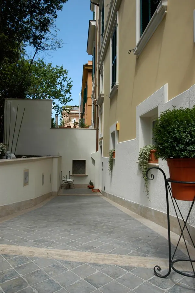 A tiled courtyard passage runs between a cream building and a low wall, with potted plants, a small metal chair and trees beyond.