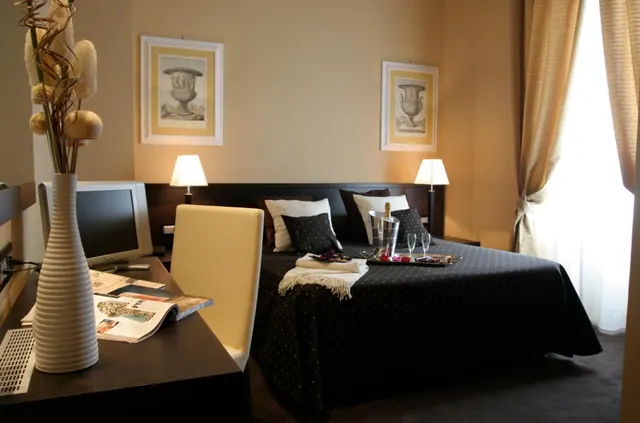 A hotel bedroom with a double bed, a tray holding a champagne bottle in an ice bucket and glasses, a desk with a monitor and a chair, and two framed prints on the wall.