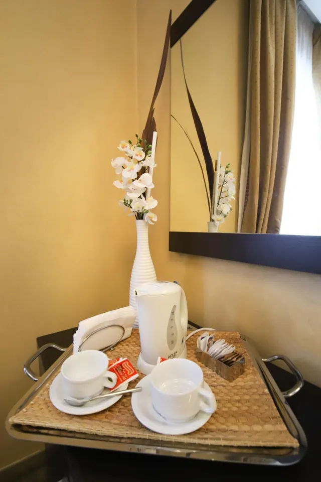 A tray on a dark surface holds an electric kettle, two cups on saucers, a basket of sugar sachets and a vase of white orchids beside a mirror.