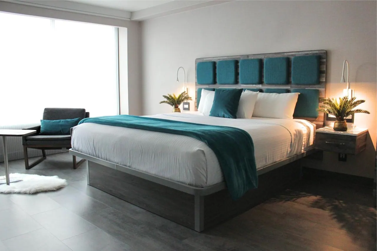 A bedroom with a king bed under a teal upholstered headboard, a teal throw, an armchair beside a large window, and potted plants on the bedside tables.