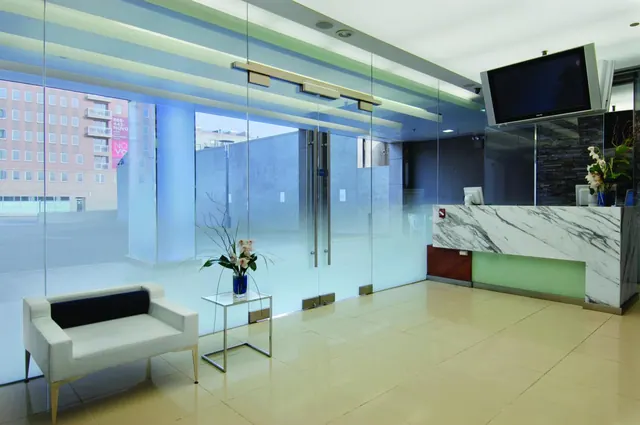 A glass-walled lobby with a white armchair, a side table with flowers, a marble reception desk and a wall-mounted television, with a brick building visible through the glass.