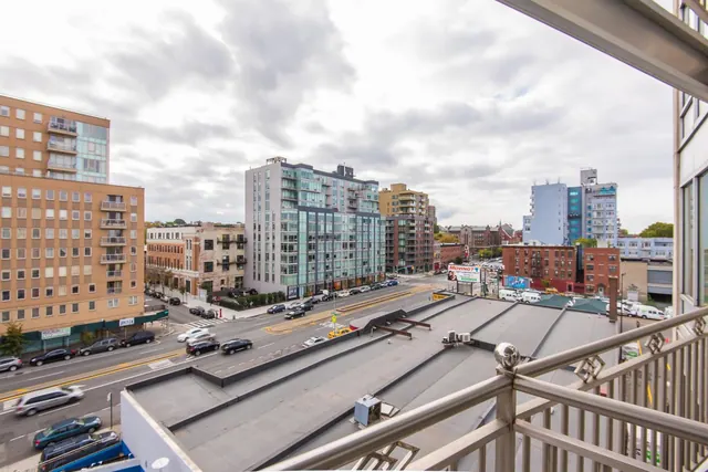 View from a balcony with a metal railing over a street with parked cars, low flat roofs and apartment buildings under an overcast sky.