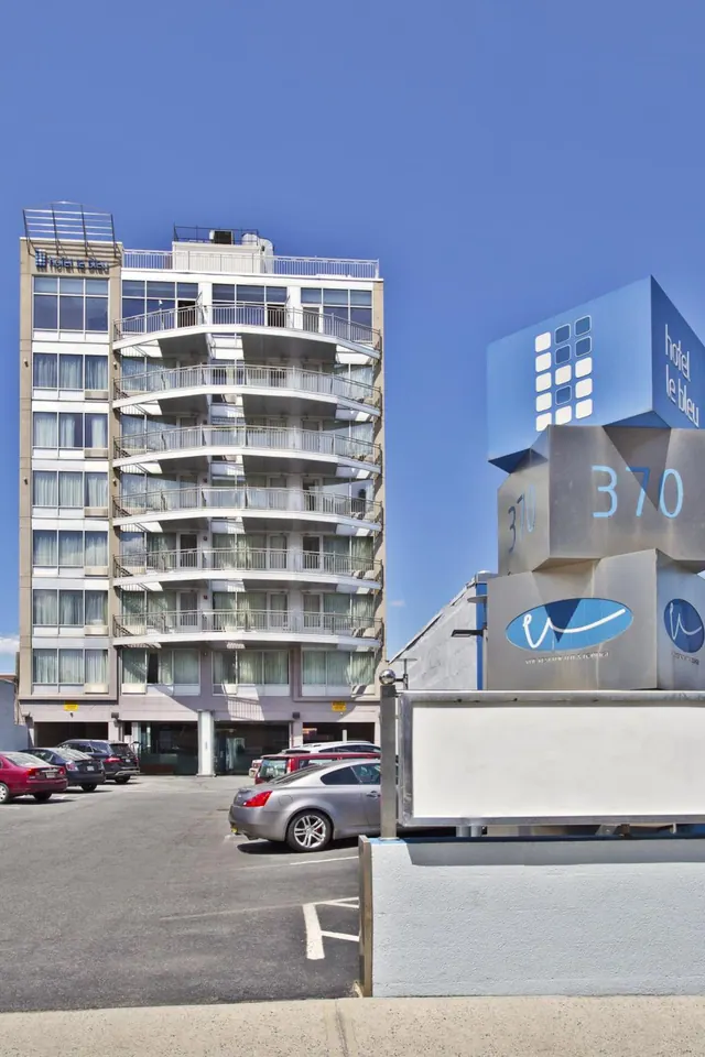 Exterior of Hotel Le Bleu, a multi-storey building with curved balconies, seen across a parking lot with parked cars and a blue and silver sign.