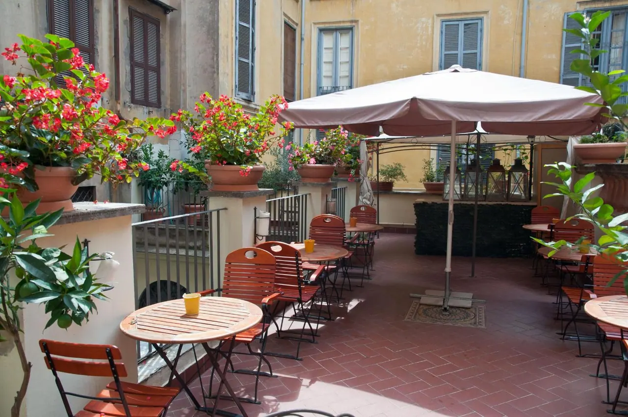 A sunlit outdoor terrace with wooden tables and folding chairs, a large parasol, potted red flowers and lanterns, facing yellow building walls and shuttered windows.