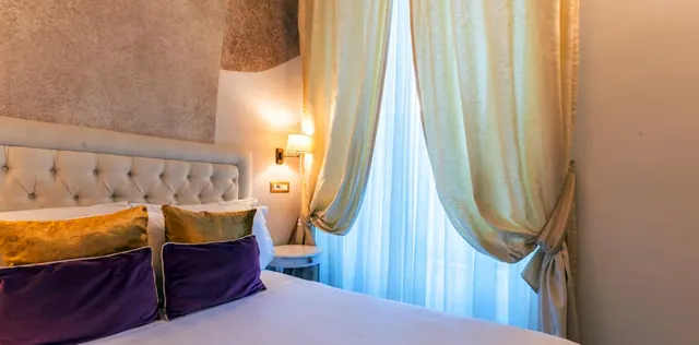 A bedroom with a tufted headboard, purple and gold cushions on the bed, a wall lamp, and sheer and drape curtains at a window.