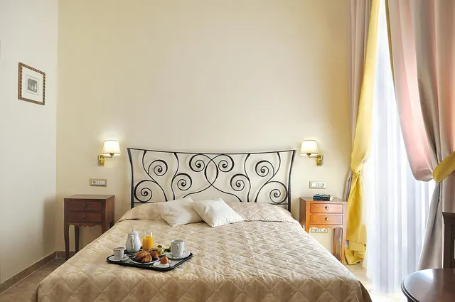 A double bed with a wrought iron headboard and a breakfast tray on the quilt, flanked by wooden bedside tables and wall lamps, with sheer curtains at a tall window on the right.
