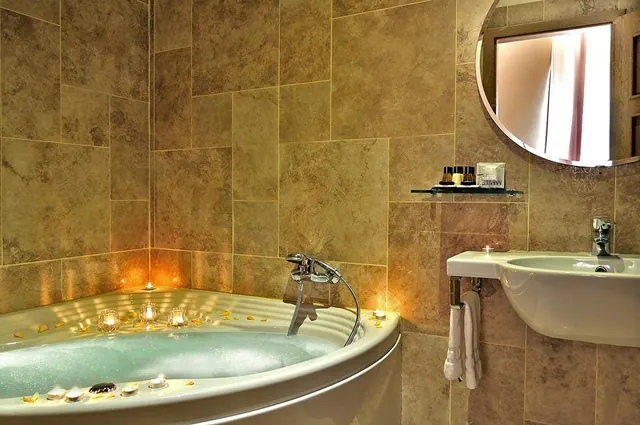 A bathroom with a corner bathtub filled with bubbling water, floating candles and petals, tiled walls, a wall-mounted sink and towels hanging on a rail.
