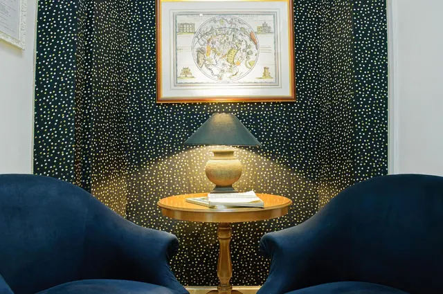 A seating corner with two navy velvet armchairs, a round wooden side table holding a lamp and books, and a framed antique map above a starry patterned curtain.