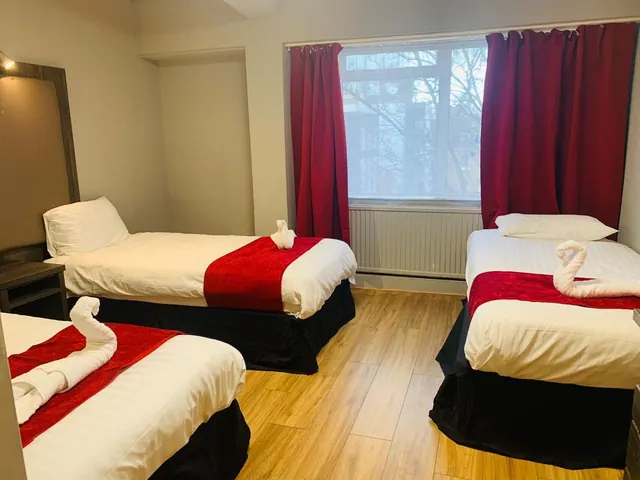 A bedroom with three single beds with white linen and red runners, red curtains, and a window showing bare trees and buildings outside.