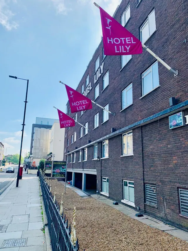 Front of a brick hotel building with three pink Hotel Lily flags on the facade, a gravel forecourt, a black railing and a street with pavement on the left.