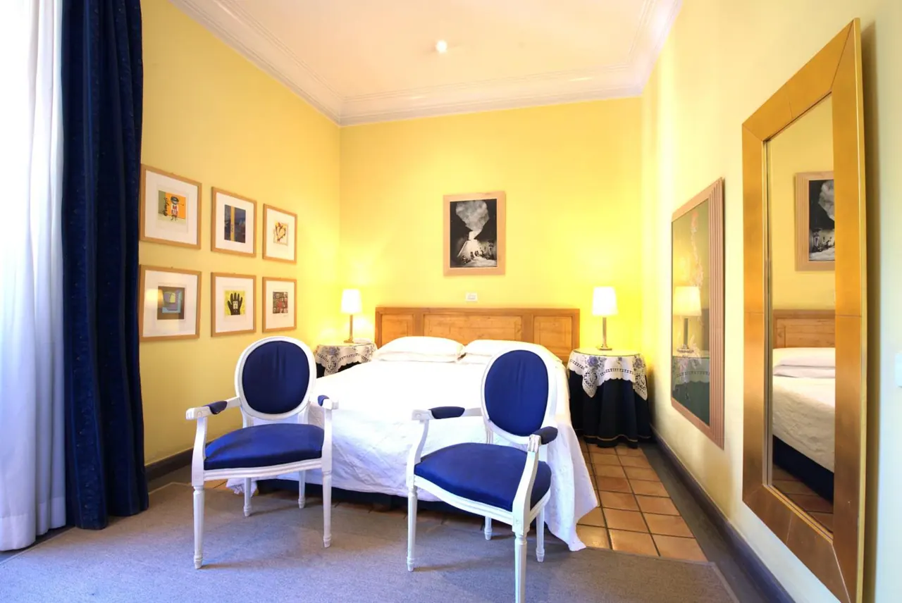 A bedroom with yellow walls, a double bed with white linens, two blue armchairs, framed artwork and a gold-framed mirror.