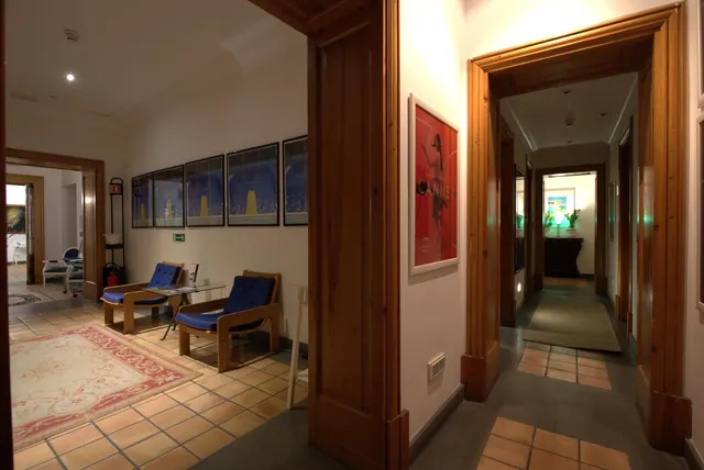 A seating area with two blue-cushioned wooden armchairs and a glass side table sits beside a corridor with framed posters, a red patterned rug, and tall wooden door frames.