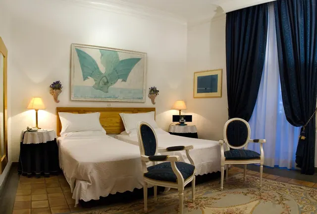 A bedroom with two beds, bedside lamps, two upholstered armchairs and navy curtains beside a tall window.