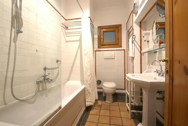 A bathroom with a bathtub and handheld shower behind a glass screen, a toilet, a pedestal sink, and terracotta floor tiles.
