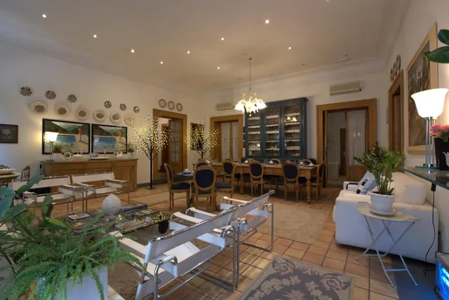 A large living and dining room with terracotta tiled floors, white leather chairs, a dining table with blue chairs, a glass-fronted blue cabinet and a chandelier.