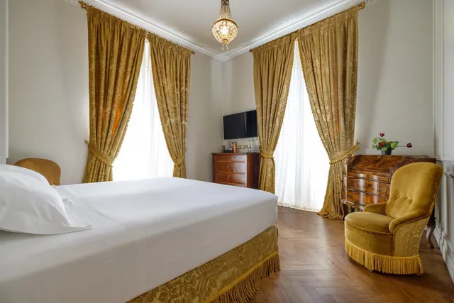 A bedroom with a large white bed, gold damask curtains, a crystal chandelier, a flat-screen television on a wooden chest, and a gold velvet armchair on a parquet floor.