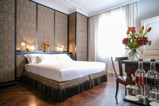 A double bed with white linens sits against a patterned wall beside a window, and a wooden table holds a vase of flowers and water bottles.