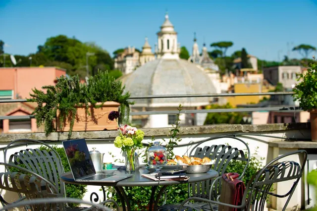 A set table with a laptop, flowers, pastries and a coffee cup on a rooftop terrace, with domed churches and buildings of the city visible beyond the railing.