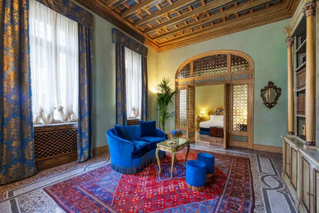 A living area with a blue velvet sofa, two blue ottomans and a glass-topped table on a red patterned rug, with an arched doorway leading to a bedroom.
