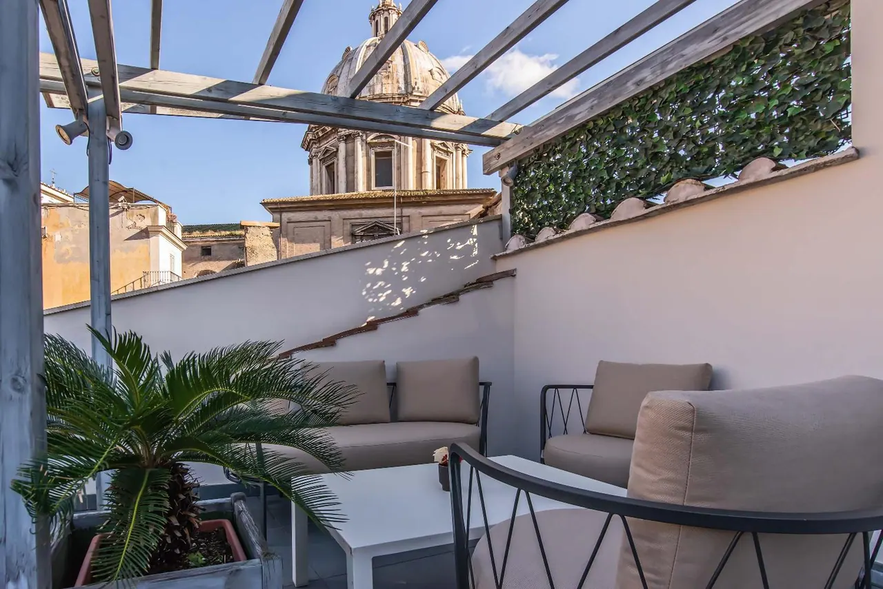 A furnished rooftop terrace with a pergola, sofa, armchairs, a coffee table and a palm plant, with a domed church visible beyond the wall.