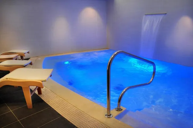 An indoor pool lit with blue light, with a chrome handrail at the steps, a water feature on the far wall, and wooden loungers with cream cushions on the left.