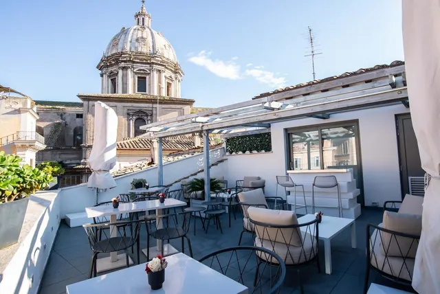 A rooftop terrace with white tables and dark metal chairs under a pergola, with a domed church and rooftops visible behind.