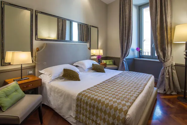 A hotel bedroom with a double bed, bedside lamps, framed mirrors, an upholstered armchair with a green cushion, and a window with long curtains.