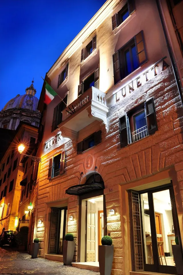 Exterior of a multi-storey hotel building at dusk with illuminated lettering on the facade, an Italian flag on a upper balcony, and a domed church visible on the left.
