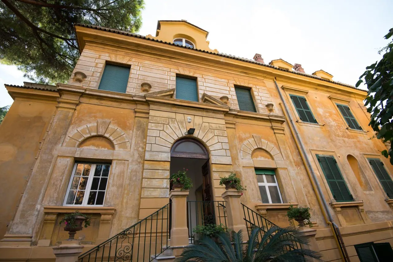 The ochre facade of a classical villa with green shutters, an arched entrance and a wrought iron staircase, photographed from a low angle.
