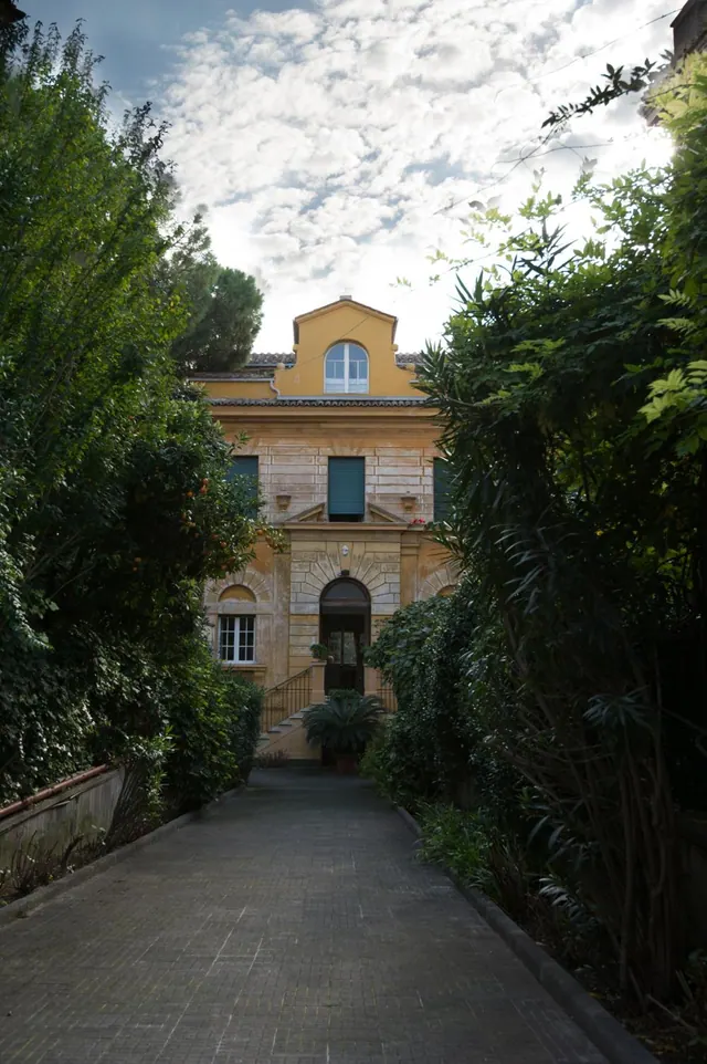 A paved path leads between dense green trees and shrubs to a yellow villa with arched windows, green shutters and an arched entrance door.
