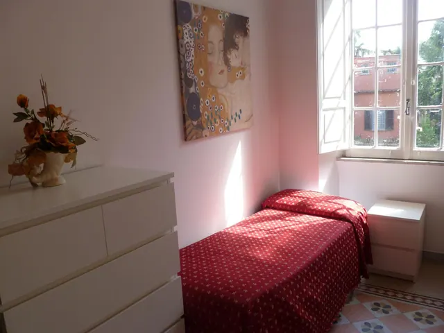 A single bed with a red patterned cover sits beside a white chest of drawers, a framed painting, and a window overlooking a brick building.