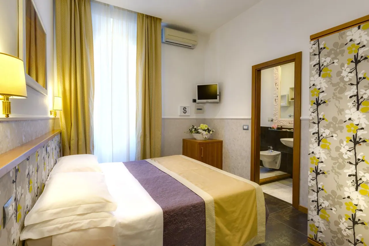 A double bed with a purple and gold throw sits in a room with a wall-mounted television and air conditioner, with an open doorway to a bathroom containing a bidet and sink.