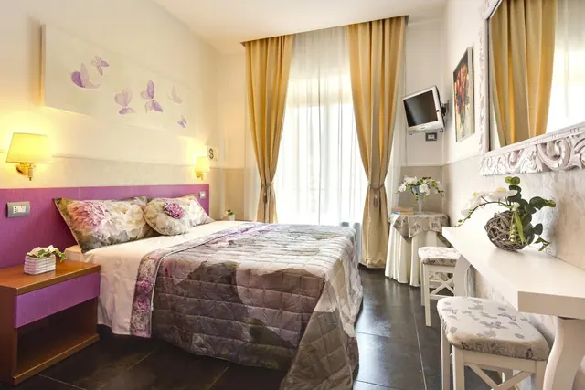 A double bed with purple floral bedding sits beside a window with gold curtains, a wall-mounted television and a white dressing table with stools.