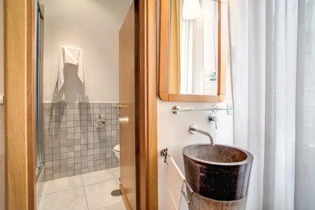 A bathroom with a glass-walled shower, grey tiled walls, a toilet, a dark stone vessel sink, and a towel on a rail beside a wooden-framed mirror.
