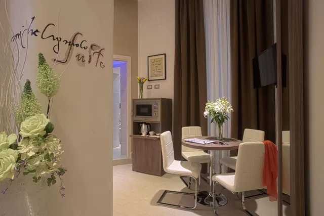 A small dining area with a round table, four white upholstered chairs, a microwave and kettle on a wooden cabinet, and a wall-mounted television.