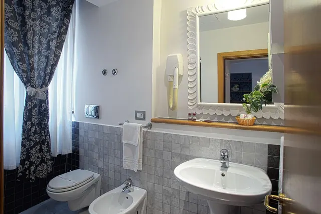 A bathroom with a wall-hung toilet, a bidet, a pedestal sink under a framed mirror, and a damask curtain beside a window.