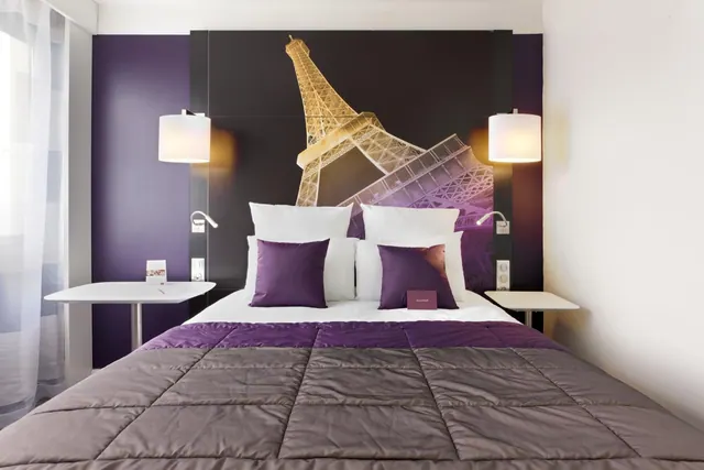 A purple-accented hotel bedroom has a double bed, bedside lamps, a small desk, and Eiffel Tower wall art.