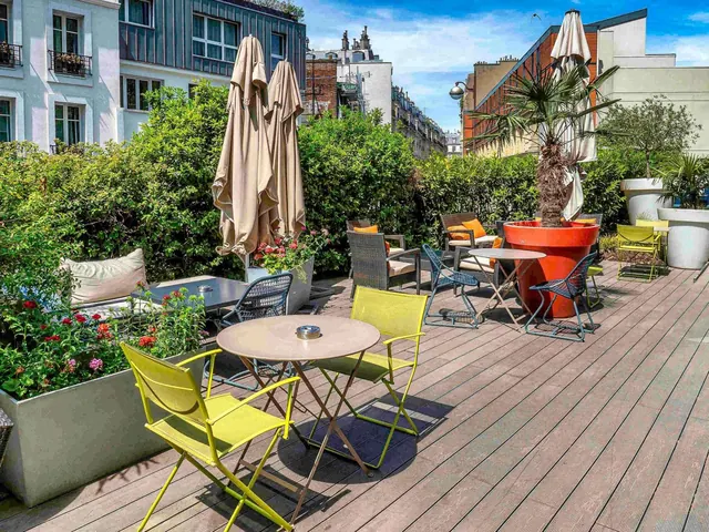 A furnished rooftop terrace with umbrellas, potted palms, plants, and views of surrounding Parisian buildings.