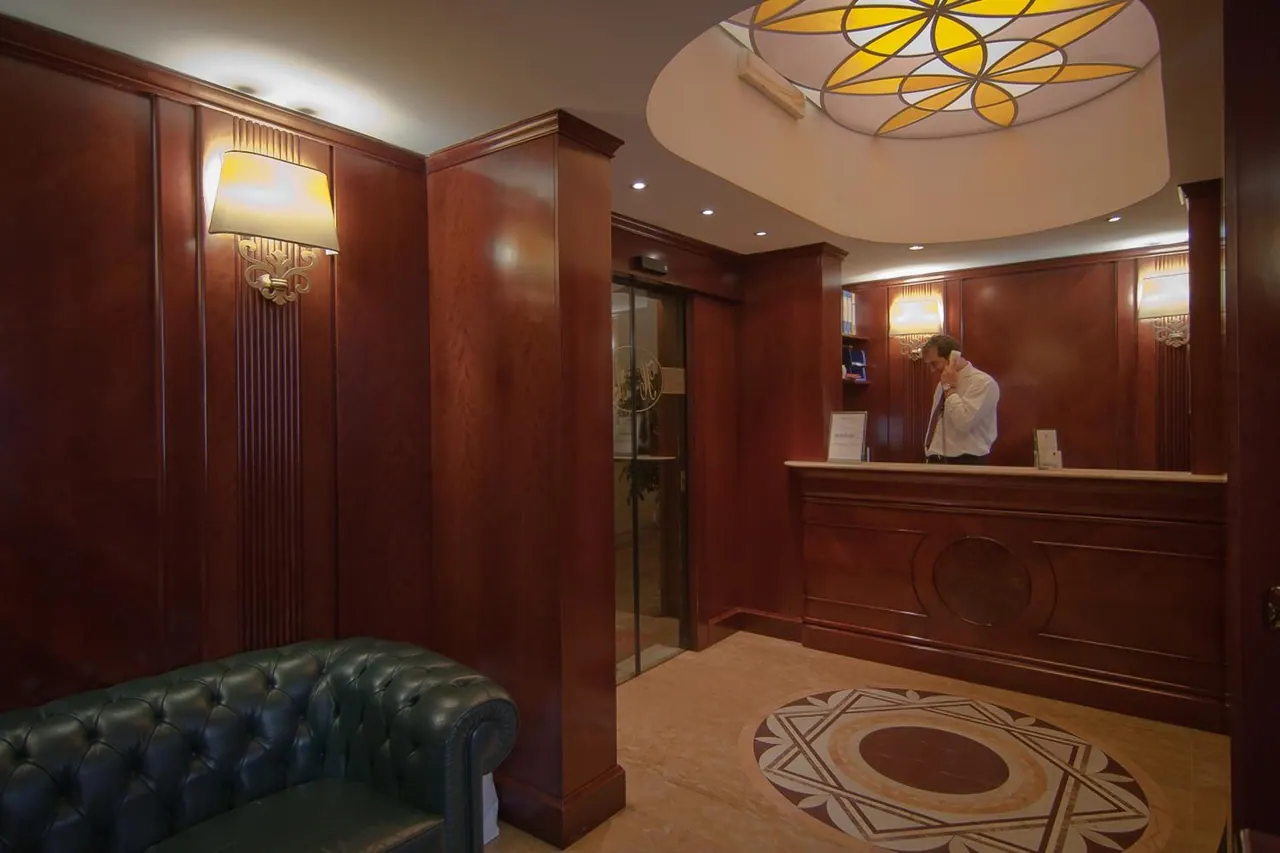 A hotel lobby with dark wood-panelled walls, a green tufted leather sofa, a reception desk with a man on the phone, glass lift doors and a stained glass ceiling dome.