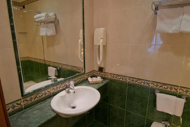 A bathroom with a white wall-mounted sink, green marble tiles, a large mirror, a wall-mounted hair dryer and folded white towels on a rail and hooks.